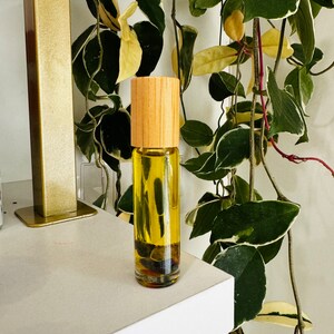 Pu&ograve; includere: Una bottiglia di vetro trasparente con un tappo di legno riempita di un liquido giallo. La bottiglia &egrave; appoggiata su uno scaffale bianco davanti a una pianta verde.