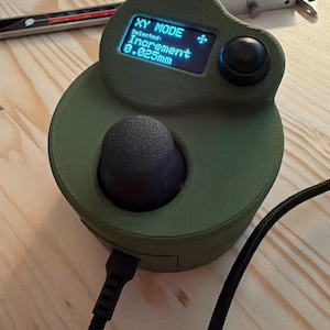 May include: An olive green electronic device with a black joystick and a small screen displaying "XY MODE" and "Increment 0.025mm." A black cable is connected to the device. A digital caliper is in the background.