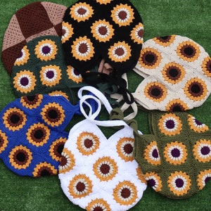 May include: A selection of handmade crochet bags and hats. The bags and hats are in various colours, including black, white, blue, and green. Each item features a sunflower design with yellow, brown, and purple accents. The items are arranged on a green surface.