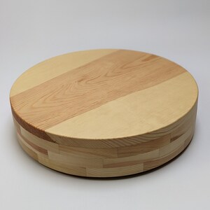 Puede incluir: Tabla de cortar o bandeja redonda de madera clara. La superficie superior presenta un patrón de vetas de madera en diagonal, mientras que los lados están construidos con piezas de madera apiladas. Los tonos de madera natural crean una estética cálida y acogedora. Diámetro aproximado de 30 cm.