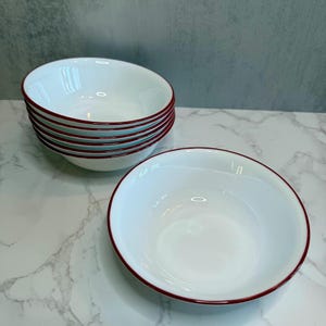May include: A stack of white ceramic bowls with a red rim. One bowl is separate from the stack. The bowls are on a white and gray marble-patterned surface.