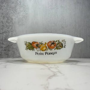 May include: White, oval-shaped casserole dish with two handles. The dish features a colorful illustration of vegetables, including tomatoes, artichokes, and onions. The words "PETITE POTAGER" are printed below the vegetables.