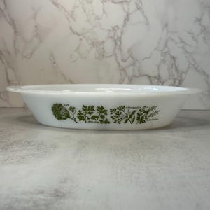 May include: A white milk glass oval baking dish featuring a green botanical print of herbs. The design includes illustrations of dill, parsley, and coriander with their names printed in small black text beneath each corresponding plant graphic.