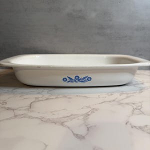 Puede incluir: Fuente rectangular blanca para hornear con bordes redondeados. Presenta un diseño floral azul en la parte delantera. Ideal para hornear y servir, mide unos 30 cm de largo.