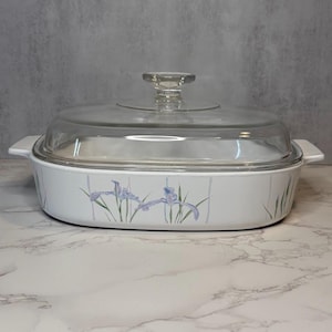 May include: White casserole dish with a clear glass lid. The dish features a floral design with blue and purple irises and green leaves. The dish has two white handles on either side.