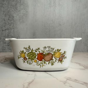 May include: White rectangular casserole dish with a colorful vegetable design. The design includes illustrations of artichokes, tomatoes, lemons, and other vegetables. The dish has two handles and the number "26" is printed on the front.