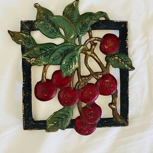 Puede incluir: Un adorno de pared de hierro fundido con un racimo de cerezas rojas con hojas verdes y detalles dorados. Las cerezas están colocadas sobre un marco cuadrado negro, creando una pieza decorativa.
