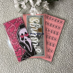 Puede incluir: Un marcapáginas rosa y negro con una ilustración de un fantasma y el texto "ALL ME!" Un sobre rosa con un billete de 100 $ dentro y la palabra "wings" escrita en él. Una hoja de papel rosa con corazones y el texto "5 €" repetido varias veces.