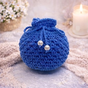 May include: A small, round, blue crocheted pouch with a drawstring closure. Two white pearl beads are attached to the front. The pouch is sitting on a textured surface, with a candle and flowers in the background.