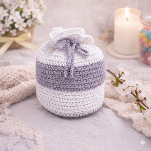 May include: A hand-crocheted pouch in white and grey, with a drawstring closure. The pouch features a white top and bottom section, with a grey band in the middle. The background includes a lit candle, flowers, and a jar of colourful sweets.