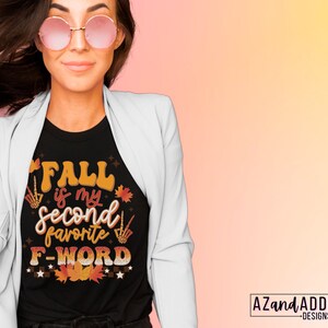 Pu&ograve; includere: T-shirt nera con la scritta "FALL is my second favorite F-WORD" in arancione, marrone e bianco. Il design include foglie autunnali e gesti delle mani di scheletro. La maglietta &egrave; indossata con un blazer bianco e occhiali da sole rotondi.