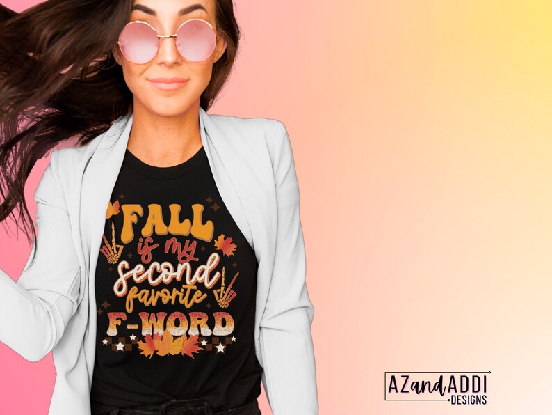 Pu&ograve; includere: T-shirt nera con la scritta "FALL is my second favorite F-WORD" in arancione, marrone e bianco. Il design include foglie autunnali e gesti delle mani di scheletro. La maglietta &egrave; indossata con un blazer bianco e occhiali da sole rotondi.