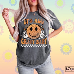 May include: A gray t-shirt with a retro-style smiley face graphic. The smiley face is wearing a leopard print pattern and has peace signs for hands. The text "It's All Gravy Baby" is printed below the smiley face.