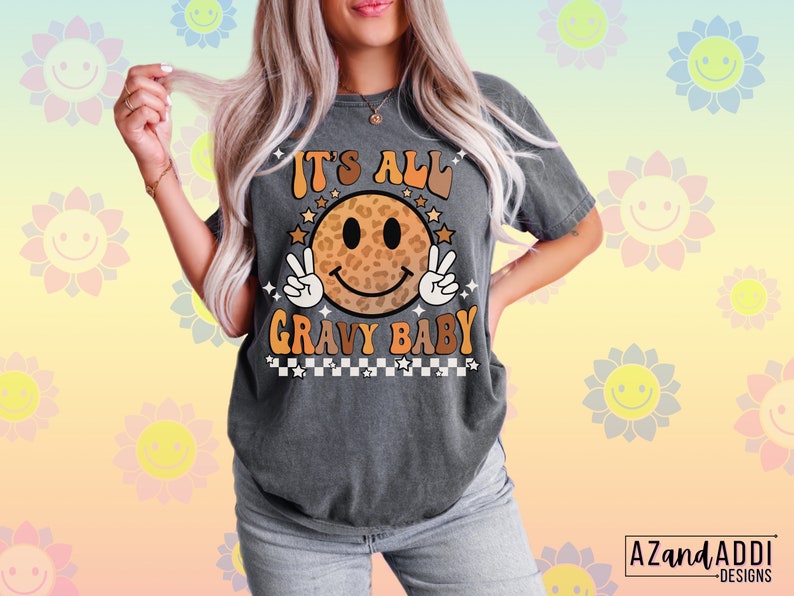 May include: A gray t-shirt with a retro-style smiley face graphic. The smiley face is wearing a leopard print pattern and has peace signs for hands. The text "It's All Gravy Baby" is printed below the smiley face.