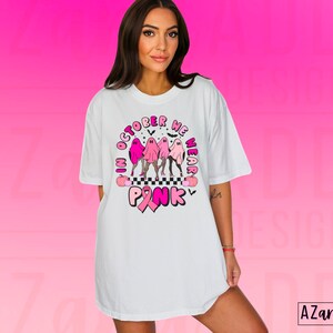 May include: White t-shirt with a graphic design featuring pink ghosts, the text "In October We Wear Pink," and a pink ribbon. The design includes a checkered pattern and bats, promoting breast cancer awareness.