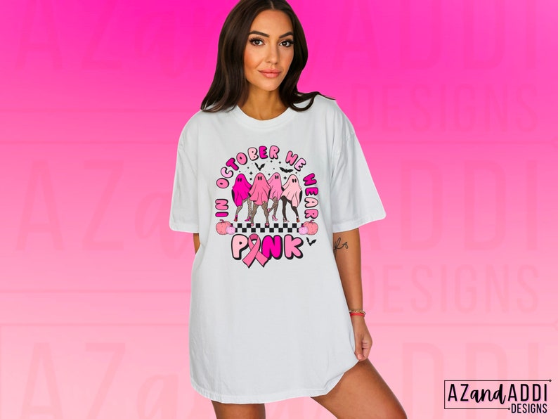 May include: White t-shirt with a graphic design featuring pink ghosts, the text "In October We Wear Pink," and a pink ribbon. The design includes a checkered pattern and bats, promoting breast cancer awareness.