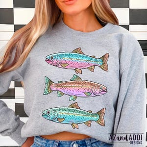 Puede incluir: Una sudadera gris con tres ilustraciones de truchas coloridas. Cada pez tiene una combinaci&oacute;n de colores &uacute;nica, incluyendo azul, rosa y marr&oacute;n, con escamas y aletas detalladas. El dise&ntilde;o incluye el texto "AZandAddi Designs" en la esquina inferior derecha.