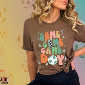 May include: A brown t-shirt with the words "GAME GAME GAME DAY" in colorful, retro-style lettering. The design includes a soccer ball, stars, and checkered flag accents. The shirt is a short-sleeved crew neck.