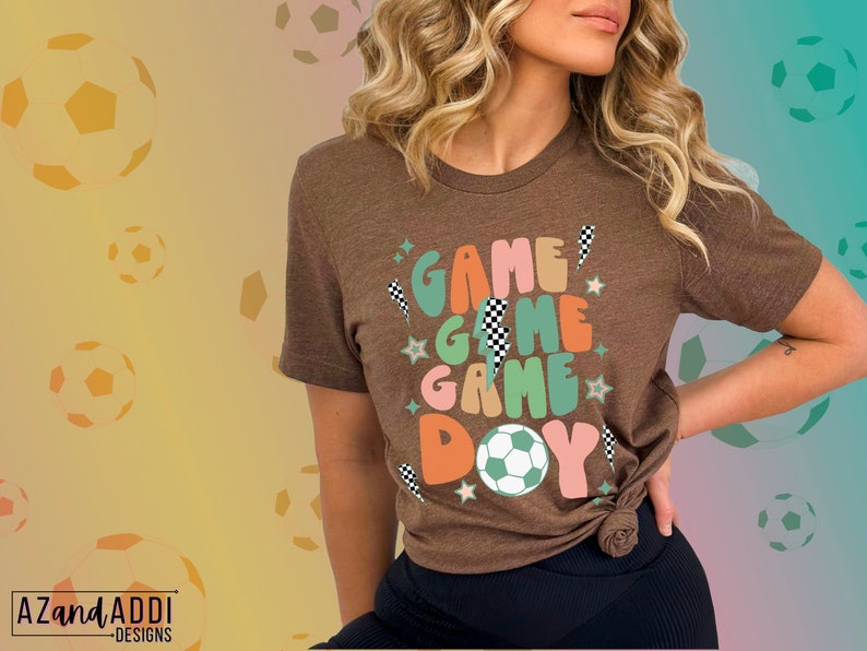 May include: A brown t-shirt with the words "GAME GAME GAME DAY" in colorful, retro-style lettering. The design includes a soccer ball, stars, and checkered flag accents. The shirt is a short-sleeved crew neck.