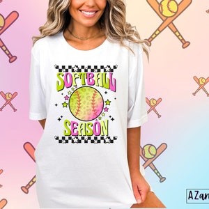 May include: White t-shirt with a graphic design of a softball with a pink and green ombre effect. The graphic also includes the text "Softball Season" with a checkered background and stars.