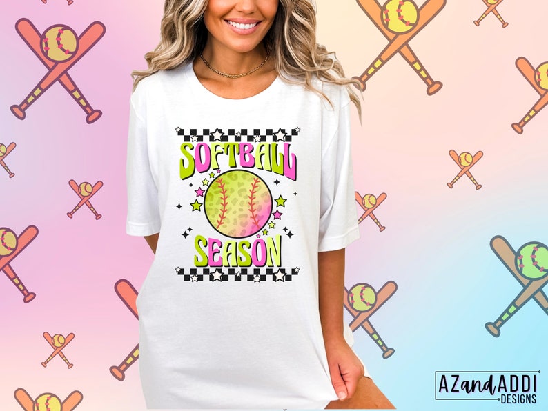 May include: White t-shirt with a graphic design of a softball with a pink and green ombre effect. The graphic also includes the text "Softball Season" with a checkered background and stars.