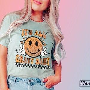 May include: A light blue t-shirt with a smiling face graphic. The face is a leopard print pattern with a peace sign on each side. The text "It's All Gravy Baby" is written in a retro font with stars and a checkered pattern.