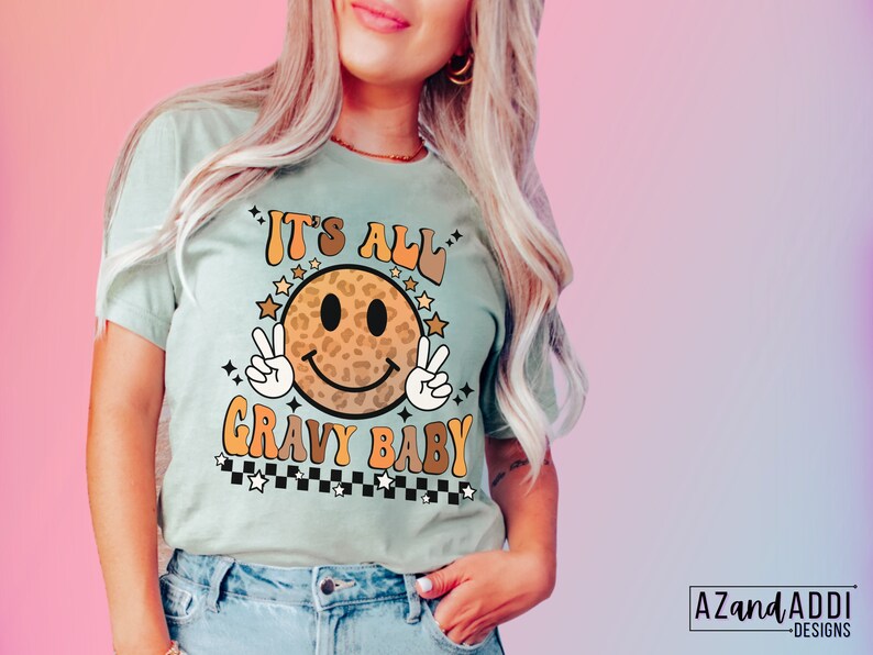 May include: A light blue t-shirt with a smiling face graphic. The face is a leopard print pattern with a peace sign on each side. The text "It's All Gravy Baby" is written in a retro font with stars and a checkered pattern.