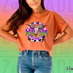 May include: Orange t-shirt with rolled sleeves, displaying a circular graphic with a bull skull and the words "Spooky Mama". The design incorporates purple, green, and Halloween motifs. The shirt is worn with blue jeans.