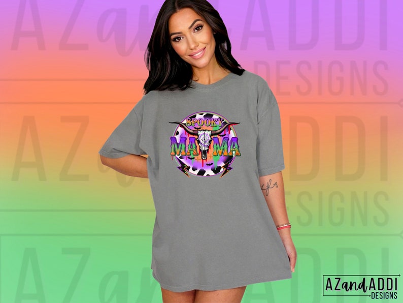 May include: A grey t-shirt with a colourful graphic that reads "Spooky Mama." The design includes a skull with horns, set against a purple, black, and white cow print. The shirt has a relaxed, oversized fit.
