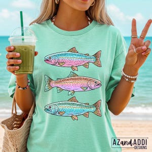 Puede incluir: Camiseta verde claro con tres ilustraciones de peces coloridos. Los peces tienen escamas azules, rosas y marrones con aletas naranjas. La camiseta tiene un corte holgado y se usa en la playa. La marca "AZANDADDI DESIGNS" est&aacute; impresa en la parte inferior derecha.
