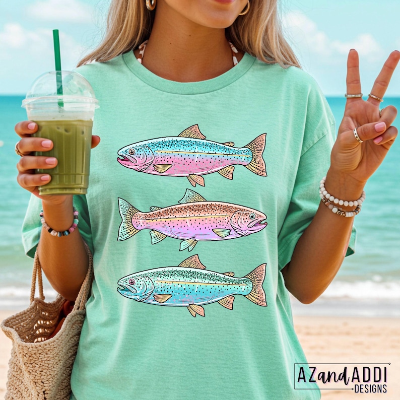 Puede incluir: Camiseta verde claro con tres ilustraciones de peces coloridos. Los peces tienen escamas azules, rosas y marrones con aletas naranjas. La camiseta tiene un corte holgado y se usa en la playa. La marca "AZANDADDI DESIGNS" est&aacute; impresa en la parte inferior derecha.
