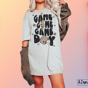 May include: A gray t-shirt with the text "Game Game Day" in black with a leopard print football helmet graphic. The text is in a retro font style with stars around it.
