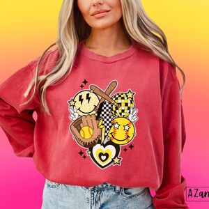 May include: A red-orange sweatshirt featuring a softball-themed graphic. The design includes smiley faces, a baseball glove, a bat, and a peace sign. The graphic has a retro, groovy aesthetic with a heart and stars.