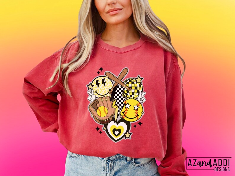 May include: A red-orange sweatshirt featuring a softball-themed graphic. The design includes smiley faces, a baseball glove, a bat, and a peace sign. The graphic has a retro, groovy aesthetic with a heart and stars.
