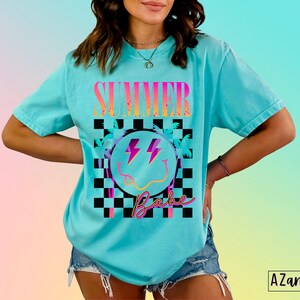 May include: A turquoise blue t-shirt with a graphic design of a smiley face with lightning bolts, palm trees, and the text "Summer Babe" in a checkered pattern.