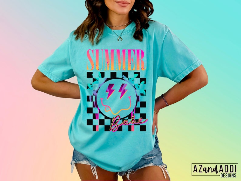 May include: A turquoise blue t-shirt with a graphic design of a smiley face with lightning bolts, palm trees, and the text "Summer Babe" in a checkered pattern.