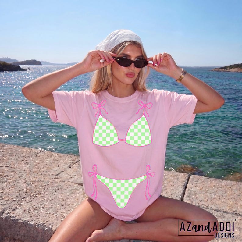 K&ouml;nnte beinhalten: Hellrosa T-Shirt mit einem Bikini-Motiv in gr&uuml;n-wei&szlig;em Karomuster. Die Konturen und Schleifen des Bikinis sind rosa. Der Text "AZAND ADDI DESIGNS" ist unten rechts im Bild zu sehen.