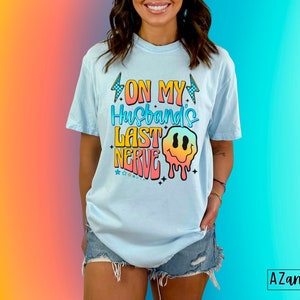 May include: A light blue t-shirt with the text "On My Husband's Last Nerve" in rainbow colors with a dripping smiley face graphic.