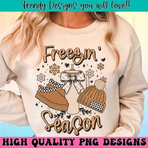 May include: A beige sweatshirt with the text "Freezin' Season" in brown lettering. The design includes a brown and white checked boot, a brown knitted hat with a white and black checked band, and a brown and white checked mug with a brown bow.