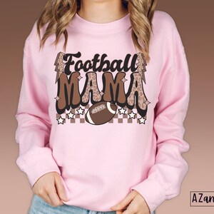 Pode incluir: Camisola cor-de-rosa com um gr&aacute;fico castanho e branco que diz "Football Mama" com uma bola de futebol e raios.