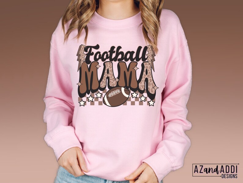 Pode incluir: Camisola cor-de-rosa com um gr&aacute;fico castanho e branco que diz "Football Mama" com uma bola de futebol e raios.