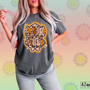May include: Dark gray t-shirt with the text "FRIENDS Giving VIBES" in a retro design. The graphic features a mix of brown, orange, and pink colors with a leopard print pattern. The shirt is a casual style.
