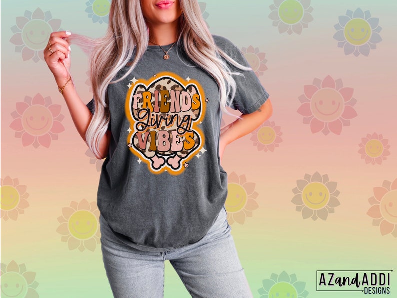 May include: Dark gray t-shirt with the text "FRIENDS Giving VIBES" in a retro design. The graphic features a mix of brown, orange, and pink colors with a leopard print pattern. The shirt is a casual style.