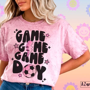 May include: Pink tie-dye t-shirt with the words "GAME DAY" in black, along with a soccer ball graphic. The design includes stars and a lightning bolt. The shirt has a crew neck and short sleeves.