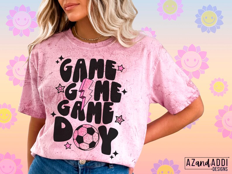 May include: Pink tie-dye t-shirt with the words "GAME DAY" in black, along with a soccer ball graphic. The design includes stars and a lightning bolt. The shirt has a crew neck and short sleeves.