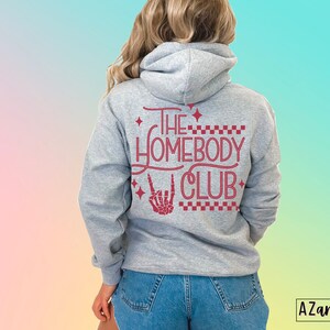 Puede incluir: Sudadera gris con capucha con el texto "The Homebody Club" en letras rojas con una mano de esqueleto haciendo un signo de rock and roll.