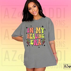 K&ouml;nnte beinhalten: Ein graues Oversize-T-Shirt mit dem Text "In My Healing Era" in bunten Retro-Lettern mit Blitzen.