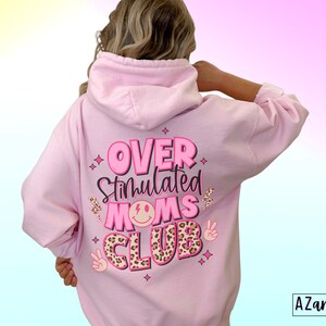 May include: Pink hoodie with the text "Over Stimulated Moms Club" in pink and black lettering with a leopard print design and peace sign graphics.