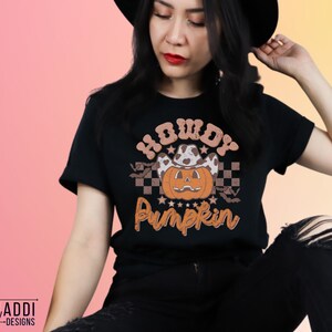 May include: Black t-shirt with a Halloween-themed graphic. The design features a pumpkin wearing a cowboy hat, the text "Howdy Pumpkin", and a checkered pattern. The shirt is a short-sleeved crew neck.