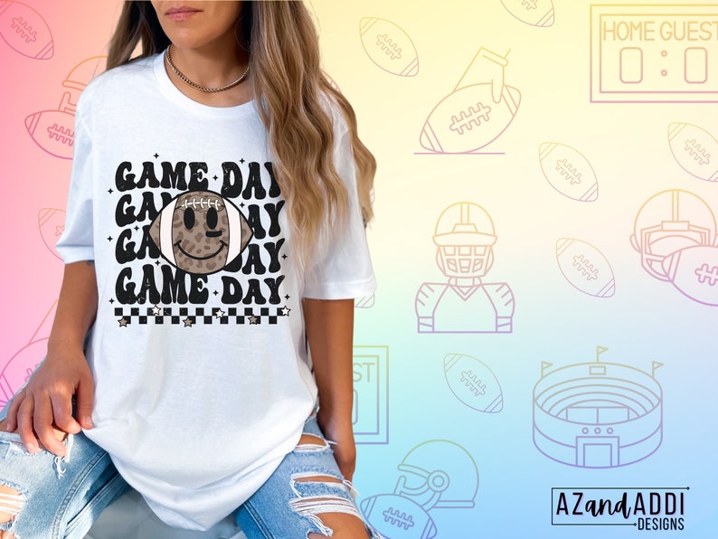 May include: White t-shirt with the text "GAME DAY" in a retro font, featuring a football with a smiley face. The design includes a black and white checkered pattern and stars. The background has football-themed illustrations.